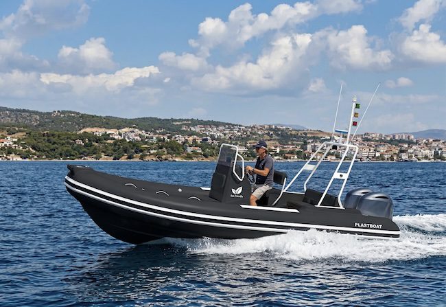 Gemini şunu dedi: An ultra-realistic photograph of a sleek, matte black RIB pleasure boat speeding through a bright blue sea, creating a white foamy wake. The boat features white horizontal stripes along its hull, a central console with a white equipment tower, and the brand name 'PLASTBOAT' on the stern. A man in his 50s with grayish hair, wearing a dark blue cap and sunglasses, is at the helm. The background shows a sunny, hilly coastline with a Mediterranean-style town under a blue sky with scattered clouds.