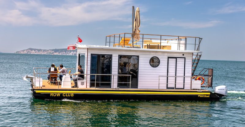 A detailed photograph of a modern, two-story houseboat with a white paneled upper structure and a black hull labeled 'ROW CLUB' in yellow. The boat is actively cruising on green-blue water, leaving a small wake. Four people are seated at a table on the front wooden deck. The rooftop terrace features wooden deck chairs, a folded sun umbrella, and railings. A small red flag is flying. In the background, a hilly coastline with buildings is visible under a partly cloudy sky. A Honda outboard motor is mounted at the stern.