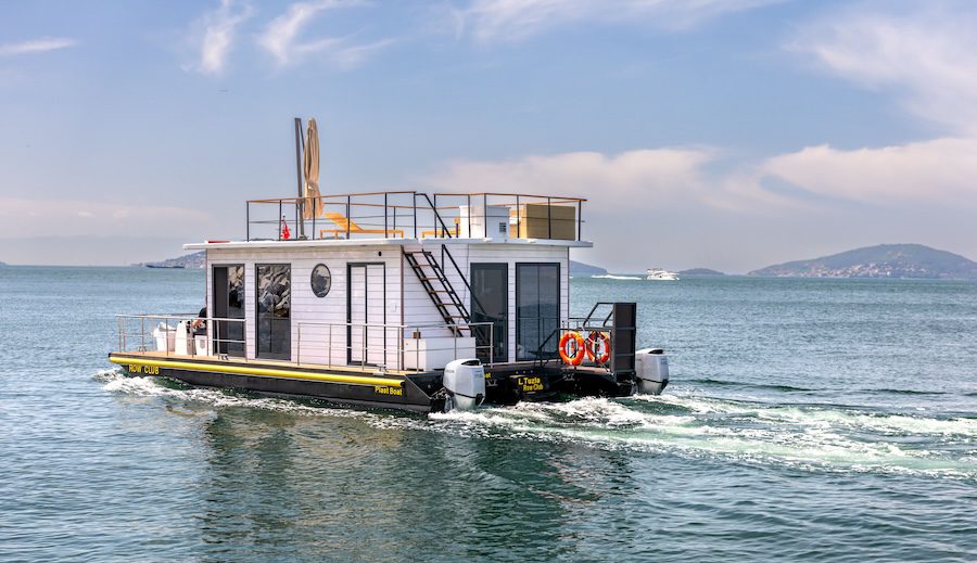 A modern white houseboat branded with 'ROW CLUB' and 'Plast Boat' cruising on a calm sea. The vessel features a top-deck lounge area, twin outboard motors, and a scenic island background under a clear sky.