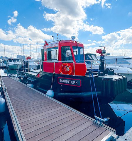 A close-up shot of a Marinturk firefighting boat moored at a dock. The vessel has a bright red cabin with "FIRE RESCUE" written on it, a black hull, and a red water cannon mounted on the bow. Other boats and a blue sky with white clouds are in the background.