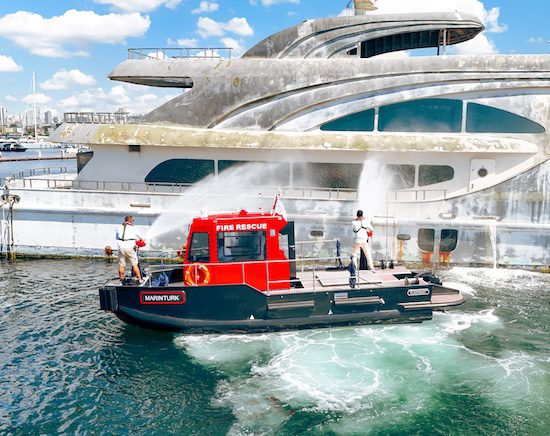 A red Marinturk fire rescue boat spraying water cannons onto a large yacht in a harbor.