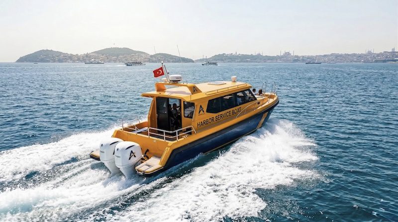 A yellow and dark blue HDPE Harbor Service Boat cutting through choppy waters at high speed, leaving a strong white wake behind. The vessel features twin outboard motors, a enclosed wheelhouse, and a Turkish flag flying from the stern. A crew member is visible inside the cabin. In the background, the iconic Istanbul skyline is visible including the Princes' Islands to the left and the silhouette of the Hagia Sophia on the right, with several ferries and cargo vessels crossing the Bosphorus.