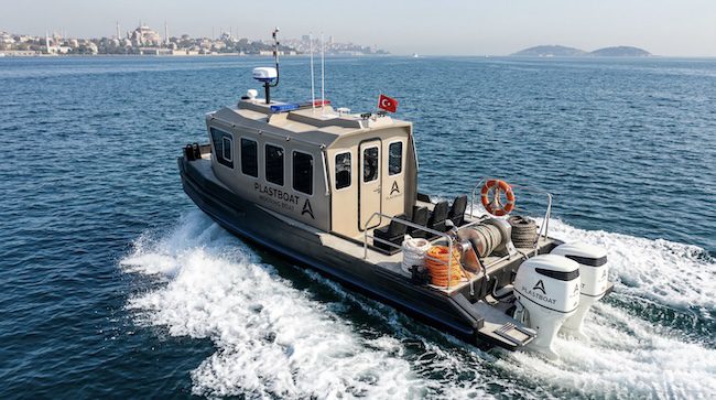 A dark grey and beige Plastboat HDPE Mooring Boat cruising at speed across the Bosphorus, leaving a white wake behind. The working deck is visibly equipped with mooring winches, coiled orange and white ropes, rubber fenders, and a bright orange life ring. A Turkish flag flies from the mast above the enclosed wheelhouse fitted with multiple windows. Twin outboard motors power the vessel from the stern. In the background, the historic Istanbul skyline is clearly visible including the silhouette of the Hagia Sophia on the left and a small island to the right.